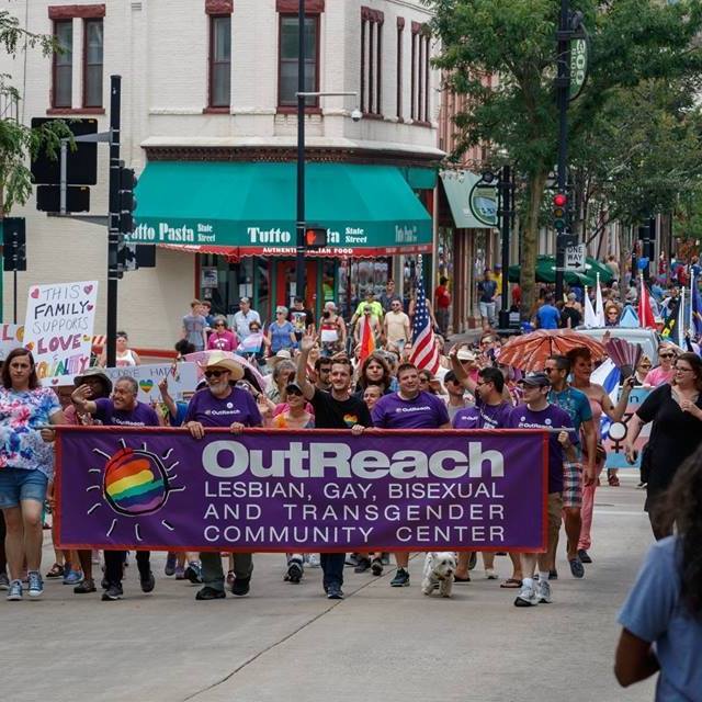 Photo of people in a parade down State Street in Madison. People at the front hold a banner that reads OutReach / Lesbian, Gay, Bisexual and Transgender Community Center