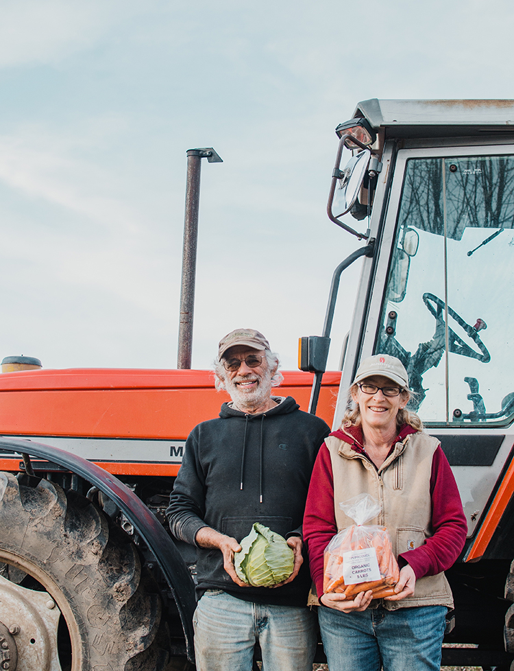 Steve Pincus holds a cabbage and Beth Kazmar holds a bag of carrots in front of their tractor.